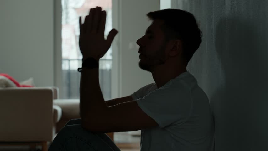 Depressed young man sitting on floor in dark room. Silhouette of young male suffering from loneliness and anxiety, covering his face with hands. Person with mental disorder