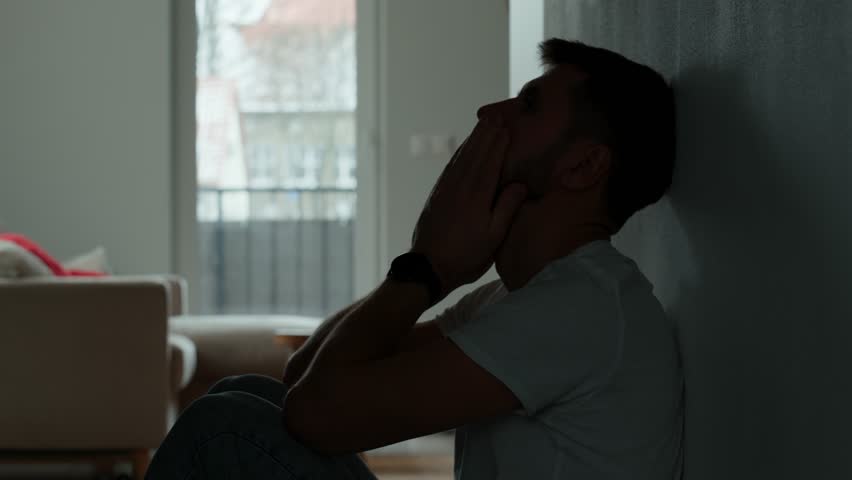 Depressed young man sitting on floor in dark room. Silhouette of young male suffering from loneliness and anxiety, covering his face with hands. Person with mental disorder