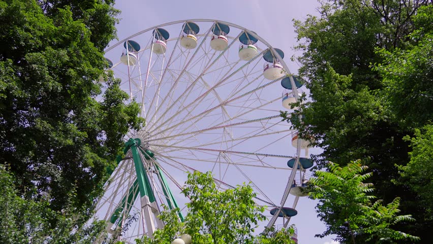 Bright Ferris wheel slowly spinning behind green trees under bright summer sun in cheerful setting of amusement park in Borjomi in Georgia.