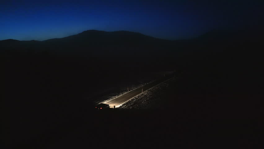 Illuminated vehicle driving on empty rural highway at night aerial view. Car with headlights riding on winter road with mountains skyline silhouette drone shot