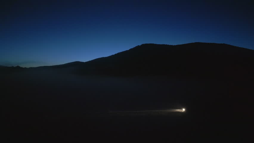 Car with headlights on road with mountains silhouette side tracking shot. Illuminated vehicle traveling at night aerial view