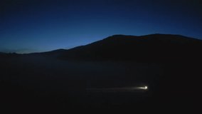 Car with headlights on road with mountains silhouette side tracking shot. Illuminated vehicle traveling at night aerial view - Powered by Shutterstock - Get 15% off with code: PIKWIZARD15