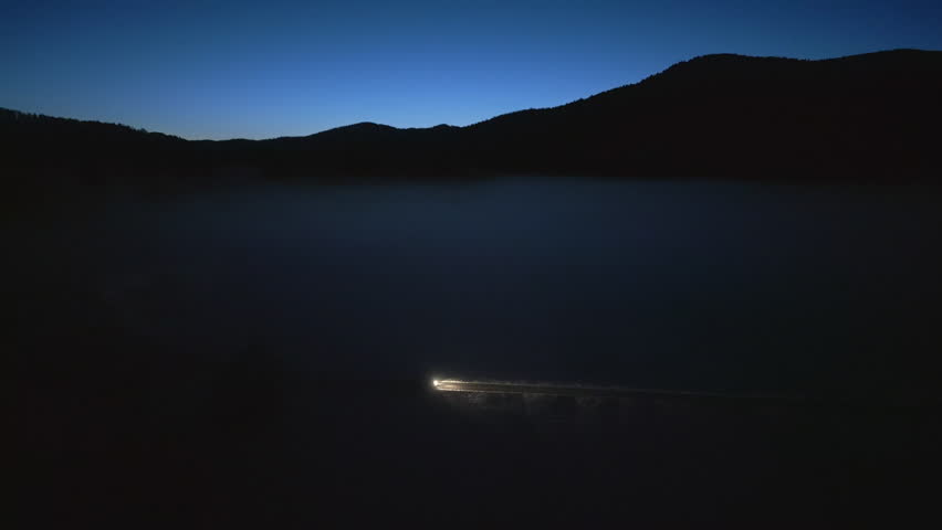 Car riding slowly on road at night with mountains skyline side tracking shot. Vehicle illuminated with headlights driving on rural highway aerial view