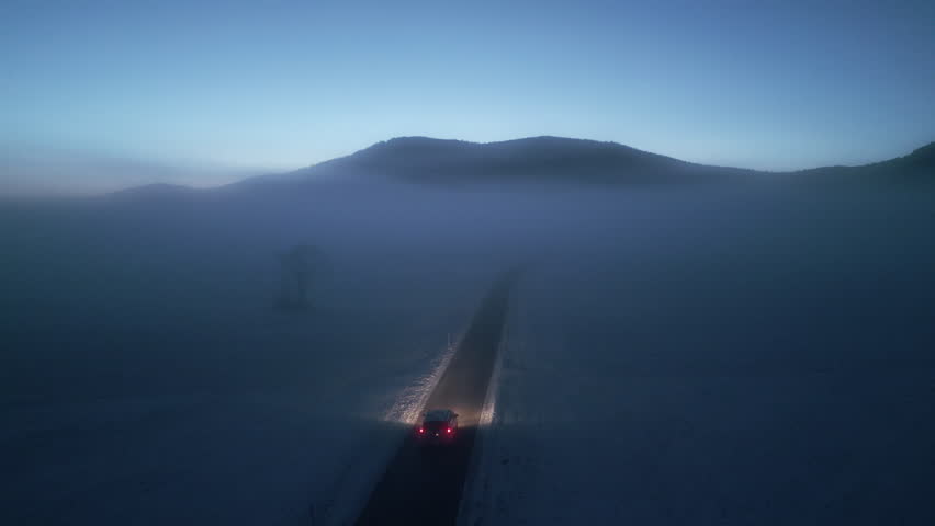 Eerie winter landscape with car on lonely road and mountains skyline aerial view. Automobile driving through morning fog on empty rural highway drone tracking shot