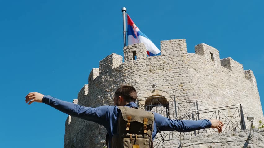 Happy man with backpack stands near the castle. A popular historical place among tourists. Old Town Fortress in Uzice, Serbia. Castle with Serbian flag on rocky mountain