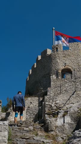 A man climbs up a stone staircase to a medieval castle. Old Town Fortress in Uzice, Serbia. Castle with Serbian flag on rocky mountain. Vertical 4k footage, Travel concept