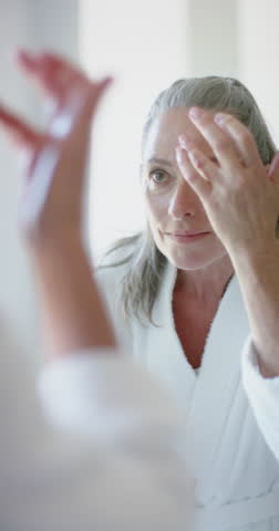 Vertical video: Applying face cream, mature woman in bathrobe caring for skin at home. Skincare, self-care, beauty, wellness, pampering, routine, slow motion