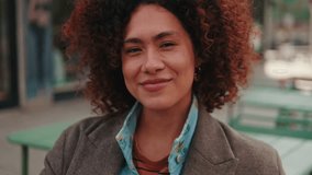 Vertical video,A young woman with curly red hair smiles at the camera outside a city cafe. She is wearing a brown jacket and a light blue and orange striped shirt. - Powered by Shutterstock - Get 15% off with code: PIKWIZARD15
