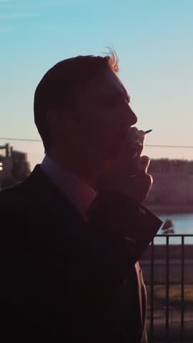 Slow motion. Sunrise silhouette of businessman smoking cigarette at the business centre roof. River and manufacture at the BG. Teal and orange. Middle steadycam shooting. Vertical Shot.