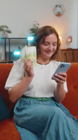 Successful rich woman holding smartphone and waving euro bill money fan sitting on sofa couch at home. Happy girl winning online lottery game planning vacation calculating budget concept. Vertical