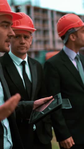 4k. UHD. Construction worker with digital pad and businessmen in suit and protective helmets talking on site about new construction. Crane and beams at the background. Teal and orange middle stabicam