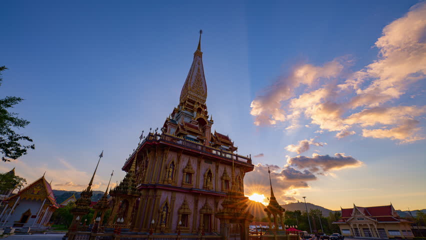 A stunning view of a traditional Thai temple wat Chalong with intricate golden architecture, captured during sunset with dramatic clouds and warm sunlight creating a serene and spiritual atmosphere.