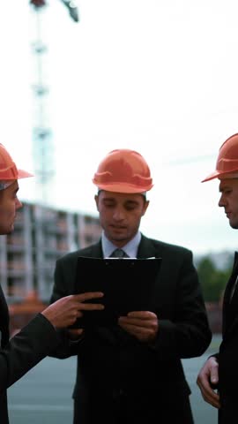 4k. UHD. Three customers: businessmen and architect sign document on text pad. They happy of a deal, smile and speaking. Orange helmets black suit and tie. Static middle shot with modern building