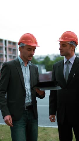 4K. ULTRA HD. Three youg successful Customers in orange helmets sign architect project on black tablet PC with newly constructed buildings at the background. Teal-and-orange middle shot. Vertical Shot