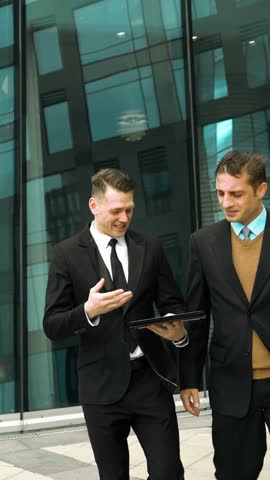 Two young successful businessmen easy communicate and discuss about something fun outdoor. Midday break. Tablet PC in hand. Glass business centre building at the background. Teal and Orange style