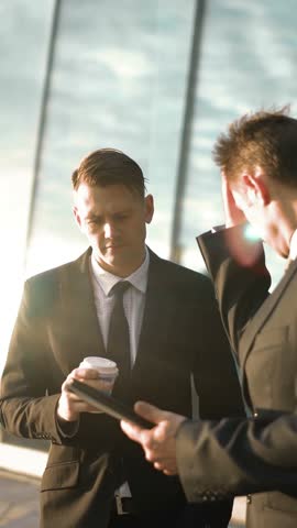 Two Young successful businessmen bidding and discuss their deal and trade. First offer his variation with tablet PC. Second debate with coffee in hand outdoor. Glass business centre building sunset at