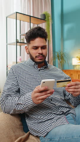 Indian man use credit bank card and smartphone while transferring money, purchases online shopping cashless order food delivery at home apartment indoors. Happy guy sits in room on couch. Vertical