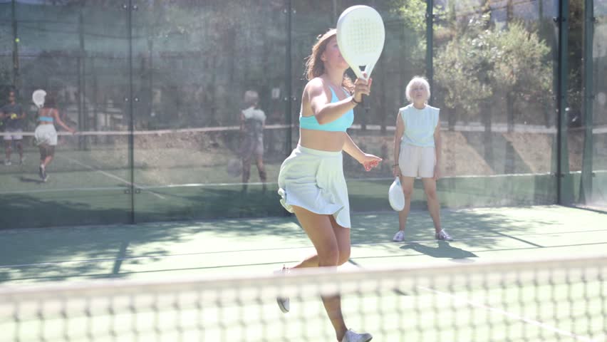 Portrait of emotional fit young woman playing padel tennis on open court in summer, swinging racket to return ball over net. Sportswoman ready to hit volley