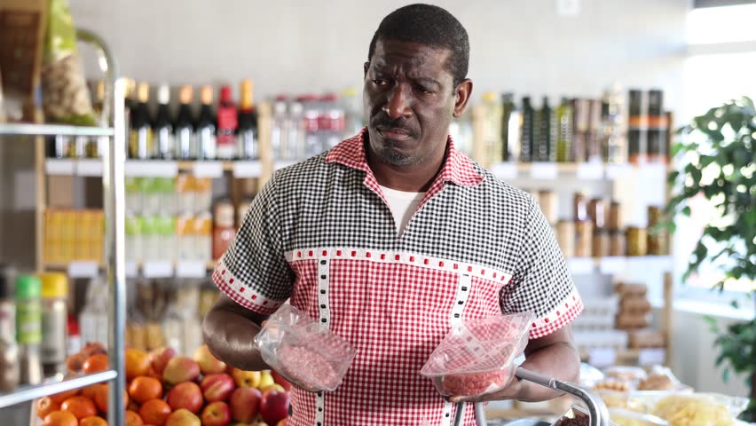 In grocery store, middle aged African man customer took out packs of raw hamburger cutlet from window, examines raw meat ware, half-finished meat for frying. 