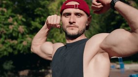 A muscular man is flexing his biceps on an outdoor sports field. He wears a black tank top and a red baseball cap. He shows off his muscles after a workout. - Powered by Shutterstock - Get 15% off with code: PIKWIZARD15