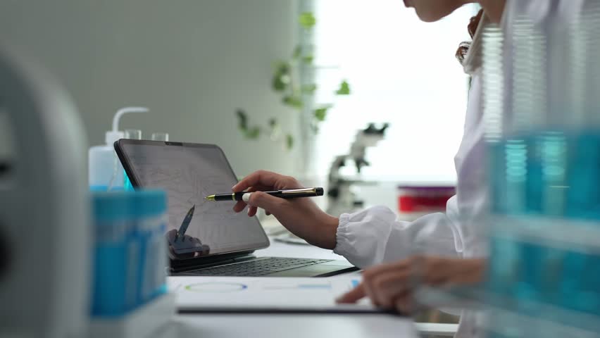 Concentrated female scientist analyzing data charts using pen while sitting at laboratory desk surrounded by test tubes and microscope - Powered by Shutterstock - Get 15% off with code: PIKWIZARD15