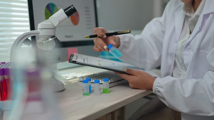 Scientist in a white lab coat conducts research, meticulously examining samples in vials and documenting observations on a clipboard, with a microscope and computer displaying data charts nearby