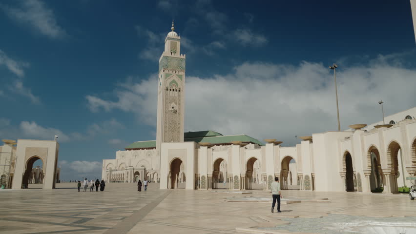 Hassan II Mosque in Casablanca, Morocco. Wide shot of people walking on a large square in front of the big white mosque. Beautiful sunny day with blue sky and white clouds.