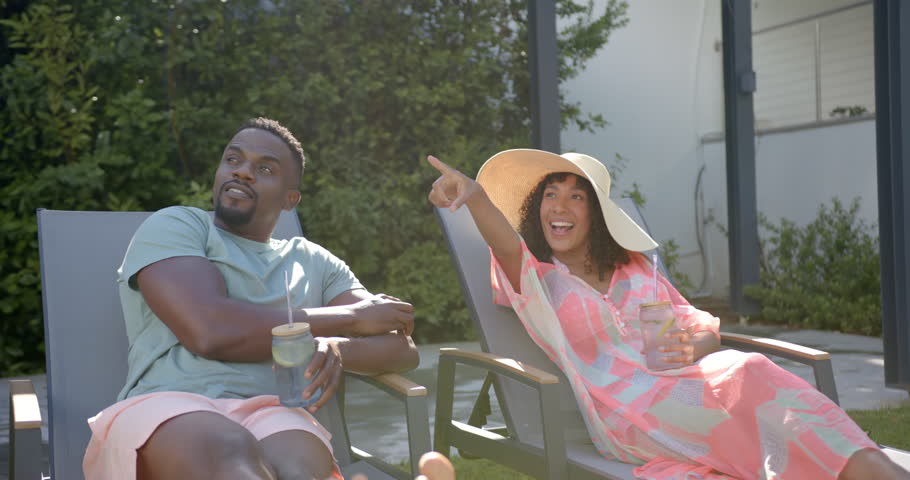 Relaxing by pool, african american couple enjoying drinks and sunshine in backyard. Vacation, leisure, relaxation, outdoors, lifestyle, enjoyment