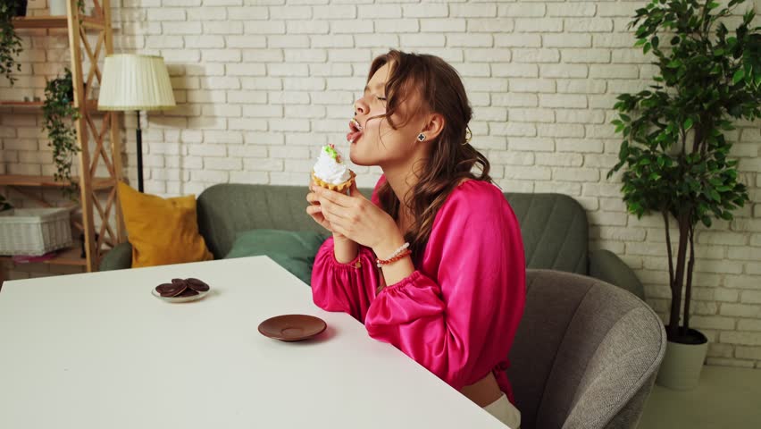 Young woman sitting at table and eating cake with whipped cream, taking a bite and chewing, enjoying the taste of sweet dessert snack, happy expression.