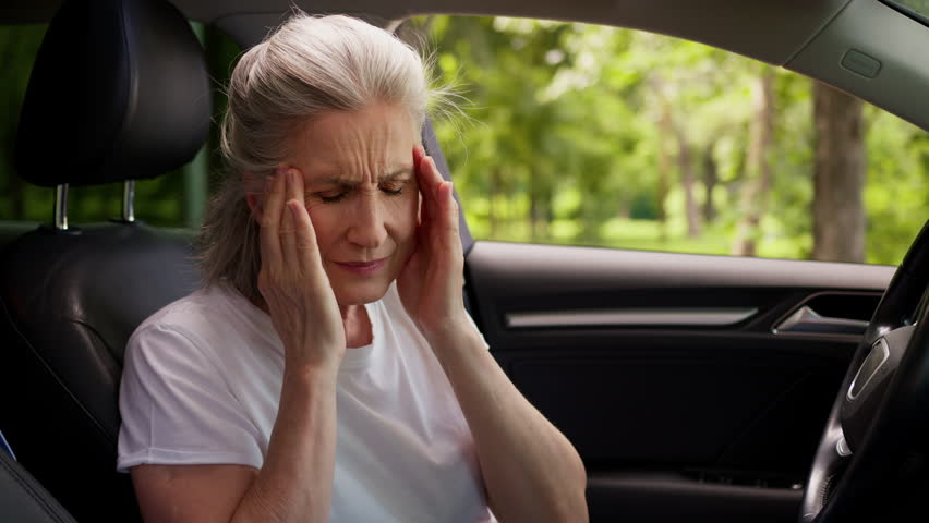 Caucasian aged mature senior woman female older automobile driver pensioner inside auto car vehicle holding head headache massaging temples pain painful tension retirement health problems at city park
