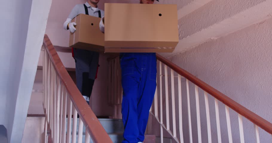 Team of young professional movers in overalls carrying packing cardboard boxes down the stairs preparing for relocating in new apartment. Move, moving day and service concept. 4k video.