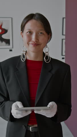 Vertical portrait shot of Asian woman smiling for camera, holding digital tablet at abstract art exhibition in gallery