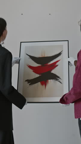 Vertical shot of female art curator and her assistant putting abstract painting on wall for exhibition in modern art gallery