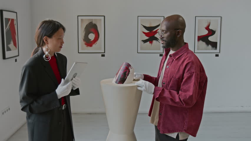 Asian art curator using digital tablet and inspecting abstract installation on pedestal, while talking to her male colleague in minimalist art gallery