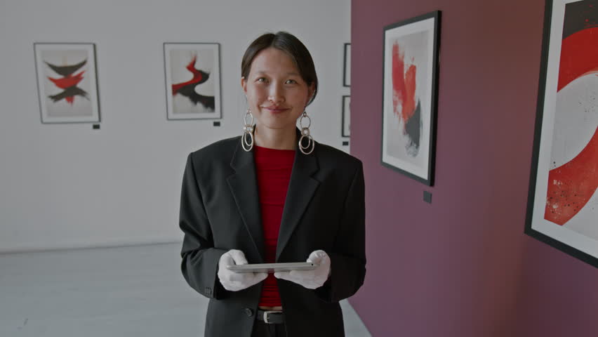 Portrait of Asian art curator, wearing smart casual clothes and white gloves, holding digital tablet, while looking at camera and smiling in minimalist art gallery