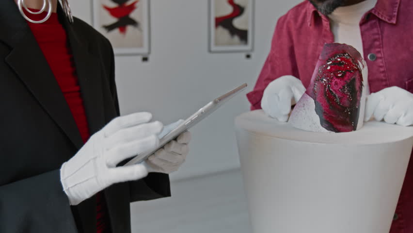 Close-up shot of unrecognizable woman using digital tablet, while her colleague checking abstract installation on pedestal in minimalist art gallery