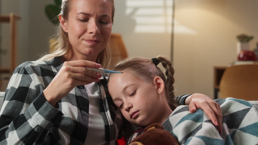 Caucasian mother mom woman worried anxiety holding thermometer high temperature fever ill sick suffering child girl daughter kid on sofa at home coronavirus symptoms health medicine problem family