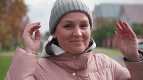 Happy woman wearing pink jacket and grey beanie adjusting beanie with a warm smile, blurred background featuring buildings and parked cars - Powered by Shutterstock - Get 15% off with code: PIKWIZARD15