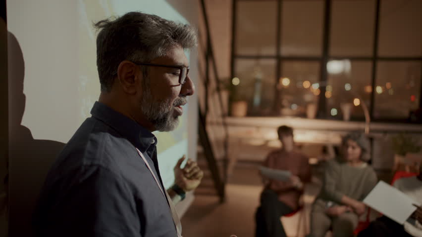 Middle-aged male speaker wearing ID badge and glasses, standing in front of projected slide and explaining concepts of energy efficient architecture during office meeting or seminar with colleagues