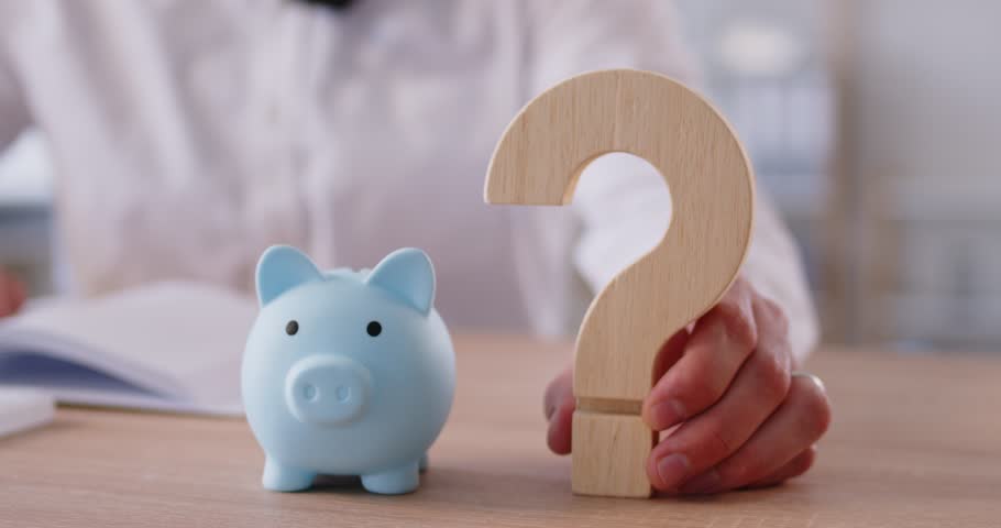Slow motion video of a businessman sitting at the desk with piggy bank and holding wooden question mark on table in office. Savings, investment and money management concept. 4k video.