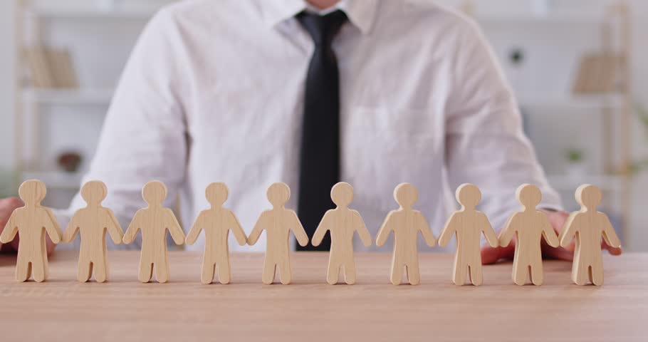 Cropped shot of young man sitting at desk, placing wooden figure of human to empty place in chain of toy people, completing line, symbolizing connection and unity. Collaboration and teamwork concept.