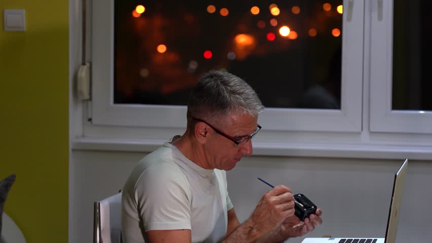 Handsome 54-Year-Old Man Spends his Free Evening, Doing his Hobby, at Home. He cleans his flash head with a fine brush. Through the window, defocused lights of moving cars and city lights are visible.