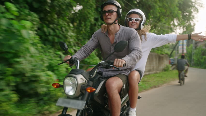 Happy couple ride on motorbike. Two lovers travel by motorcycle in tropics. Girlfriend spread arms, guy drive scooter in jungle. Travelers explore locations on bike. Man and woman enjoy adventure.