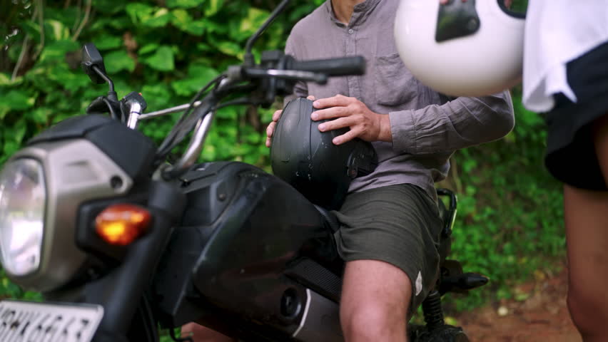 Nice happy couple puts on motorcycle helmets gets on motorbike ready to start off. People dress protection for bike, sit on scooter to go on trip in jungle. Man woman comply safety rules.