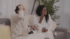 Two diverse female friends are sitting on a sofa in their living room, enjoying a relaxing evening together while applying cosmetic face masks - Powered by Shutterstock - Get 15% off with code: PIKWIZARD15