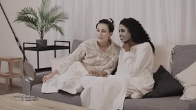 Female friends wearing bathrobes relaxing together on couch, applying purifying clay masks during home spa experience, sharing wellness moment and enjoying skincare routine - Powered by Shutterstock - Get 15% off with code: PIKWIZARD15