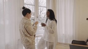 Two diverse women, wearing comfortable loungewear, stand by a window in a cozy home, sipping hot beverages from mugs and engaging in friendly conversation - Powered by Shutterstock - Get 15% off with code: PIKWIZARD15