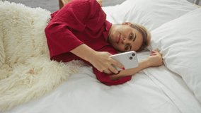 Woman relaxing on bed in cozy bedroom holding smartphone while wearing red shirt and resting amid soft pillows, creating a serene and peaceful indoor atmosphere - Powered by Shutterstock - Get 15% off with code: PIKWIZARD15