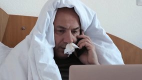 Sick middle aged man sitting on bed with laptop. He has a tissue in his nose. Person is talking on the phone, close up. Employee is working from home despite illness. Worker wrapped in warm blanket - Powered by Shutterstock - Get 15% off with code: PIKWIZARD15