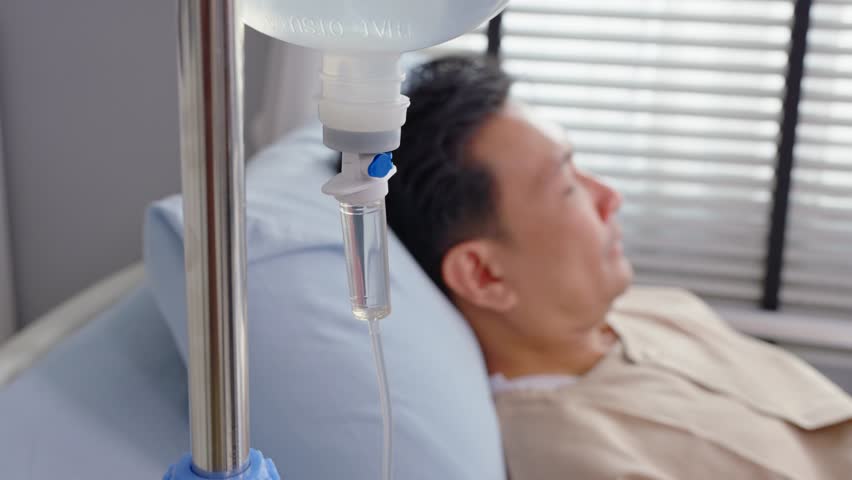 Close up of mature male patient getting saline solution in hospital ward. Senior elderly sick man lying down on bed in the recovery room waiting for treatment from medical doctor in health care clinic